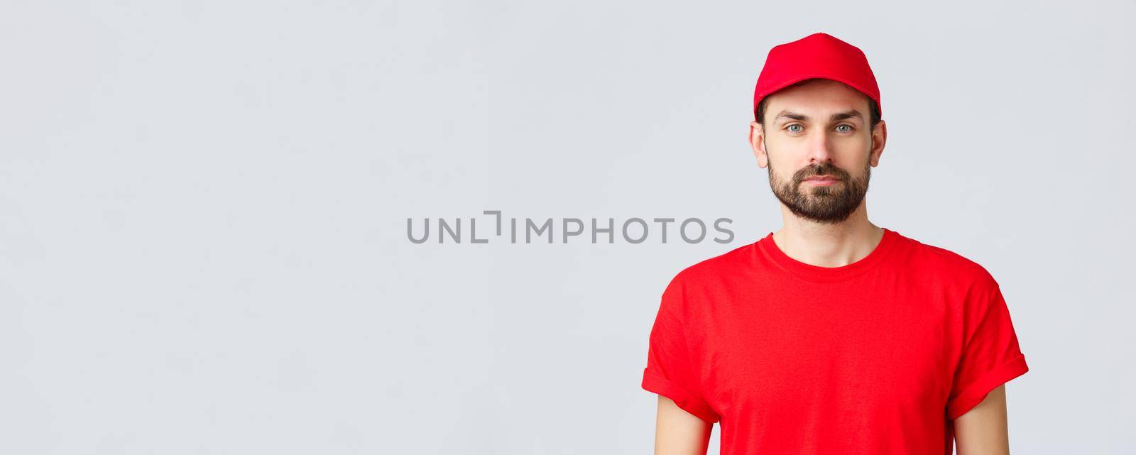 Online shopping, delivery during quarantine and takeaway concept. Young handsome bearded courier in red service uniform with cap and t-shirt, employee working transfering orders.