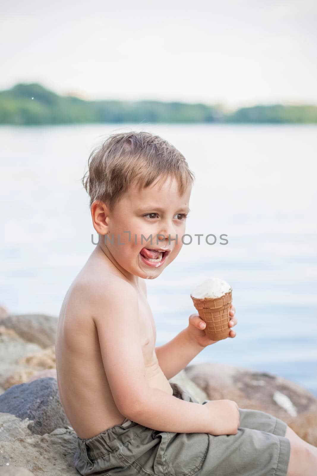 A cute blond boy appetizingly eats ice cream in the summer, sitting on the bank of the river. Cool off by the water. Funny facial expression. summer heat