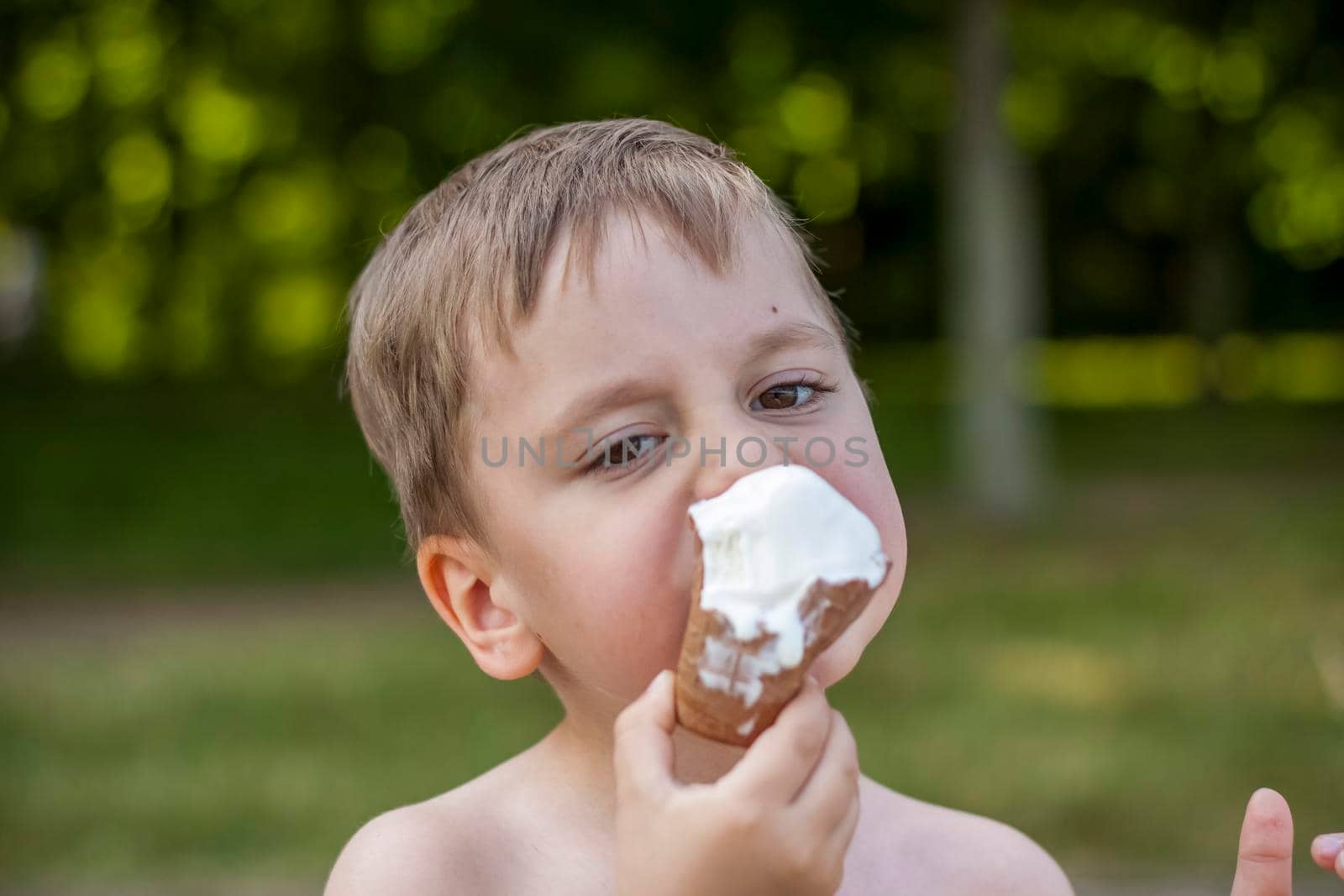 A cute blond boy appetizingly eats ice cream in the summer, sitting on the bank of the river. Cool off by the water. Funny facial expression. summer heat