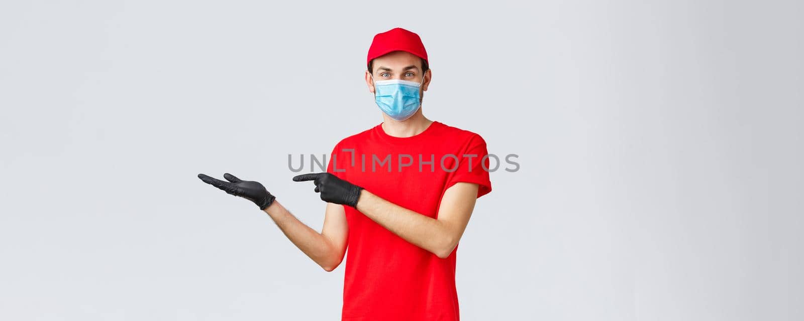 Groceries and packages delivery, covid-19, quarantine and shopping concept. Friendly courier in red uniform, face mask and gloves, introduce promo on banner, pointing left, showing advertisement.