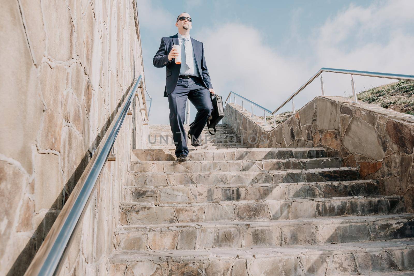 Confident middle age businessman with briefcase walking upstairs. Close-up of businessman wearing blue suit holding bag and coffee cup walking down the stairs by panophotograph