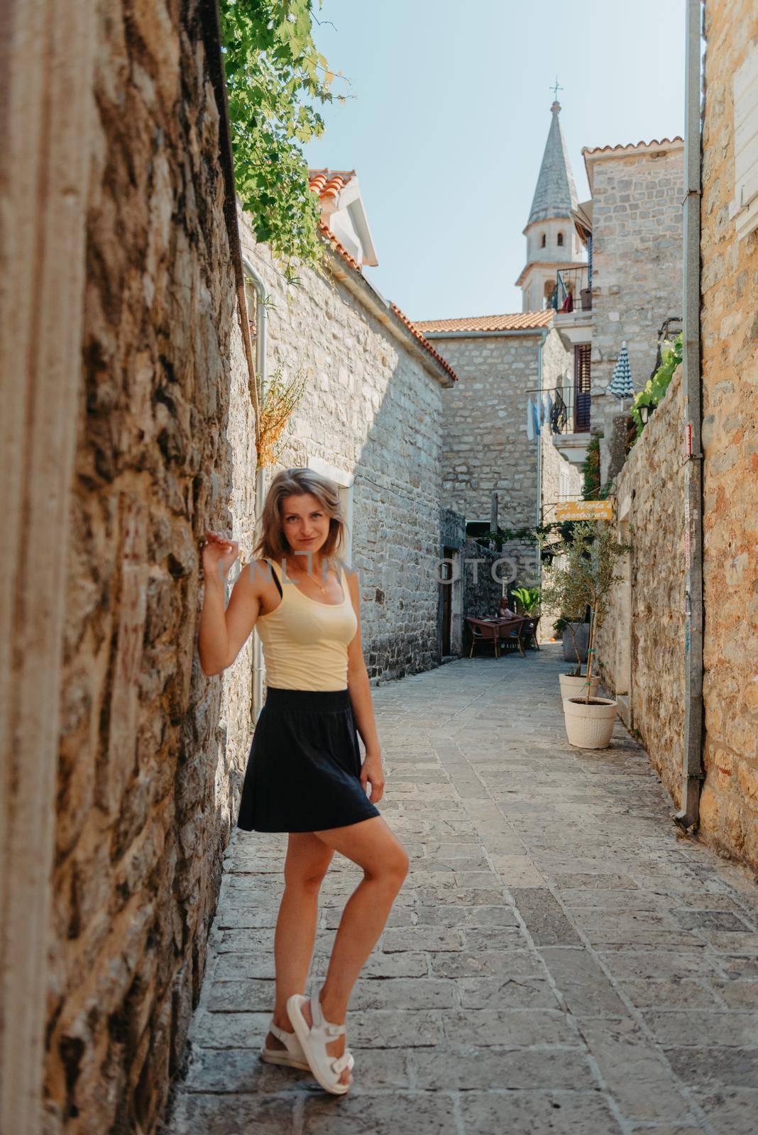 Girl tourist walking through ancient narrow street on a beautiful summer day in MEDITERRANEAN MEDIEVAL CITY , OLD TOWN bUDVA, MONTENEGRO. Young beautiful cheerful woman walking on old street at tropical town. Pretty girl looking at you and smiling