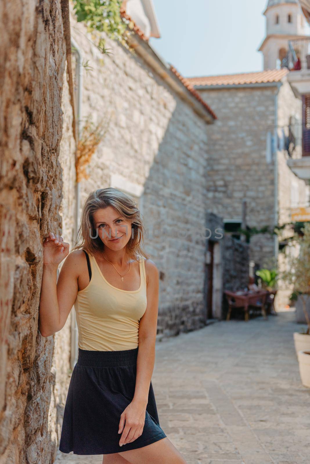 Girl tourist walking through ancient narrow street on a beautiful summer day in MEDITERRANEAN MEDIEVAL CITY , OLD TOWN bUDVA, MONTENEGRO. Young beautiful cheerful woman walking on old street at tropical town. Pretty girl looking at you and smiling