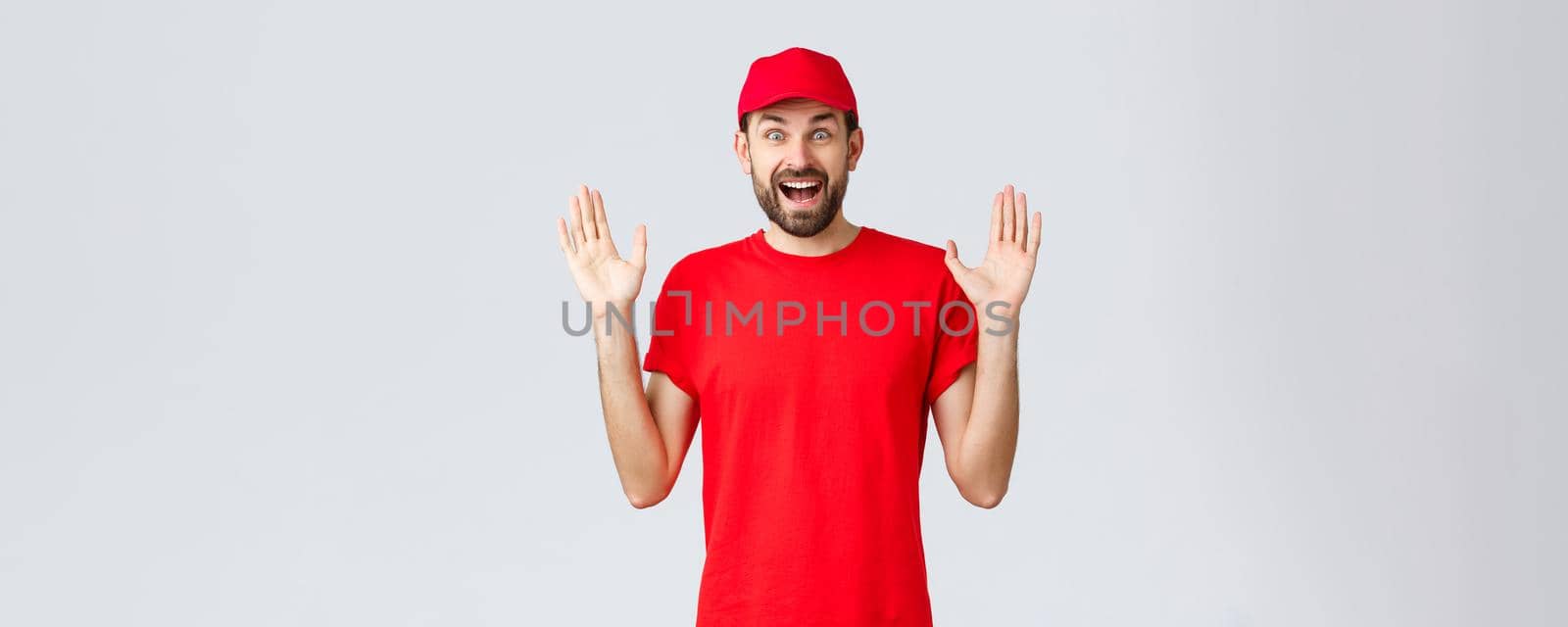 Online shopping, delivery during quarantine and takeaway concept. Happy cheerful courier in red t-shirt and cap, company uniform, hands up surprised and amused, standing grey background by Benzoix