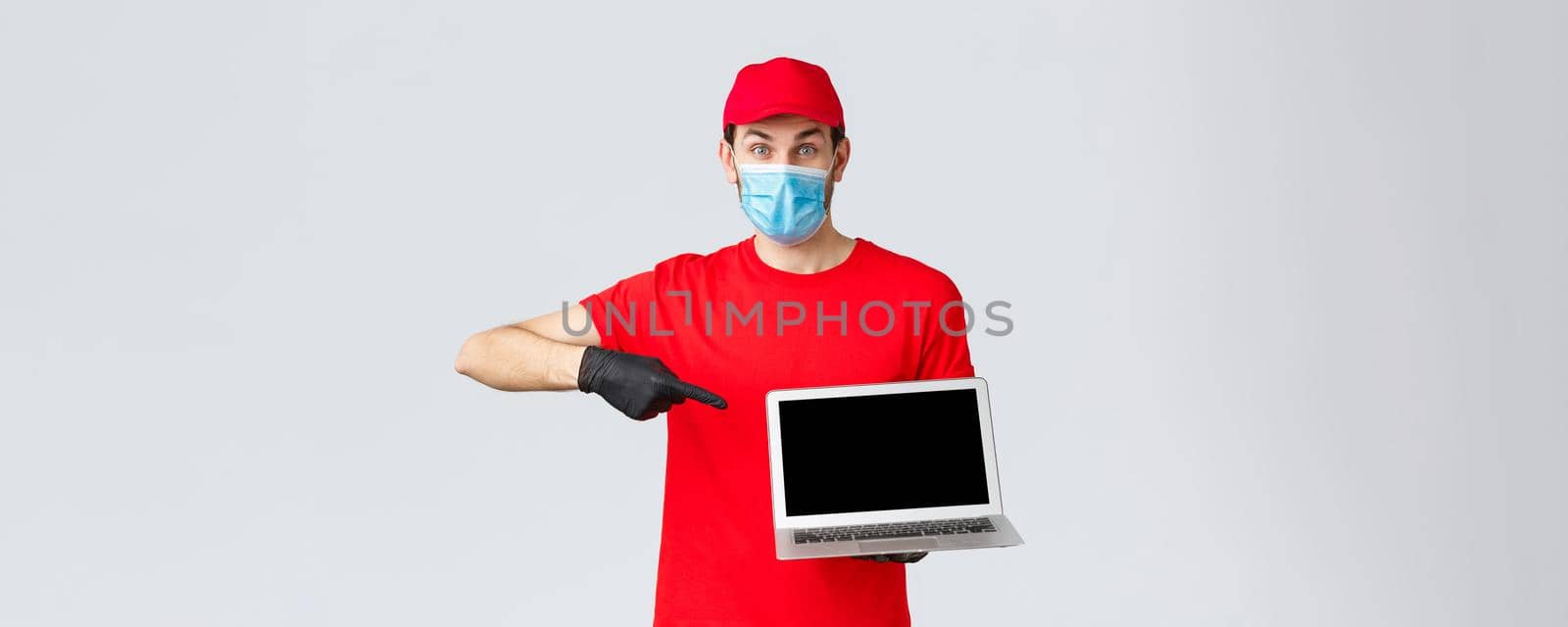 Customer support, covid-19 delivery packages, online orders processing concept. Enthusiastic courier in red uniform, gloves and face mask from coronavirus, pointing at laptop screen.