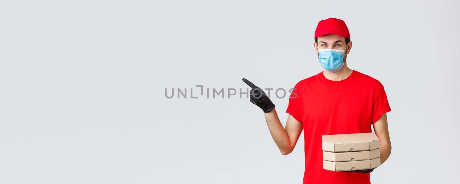 Food delivery, application, online contactless shopping and covid-19 concept. Charismatic courier in red uniform, face mask and gloves, pointing finger left, holding pizza, bring order client by Benzoix
