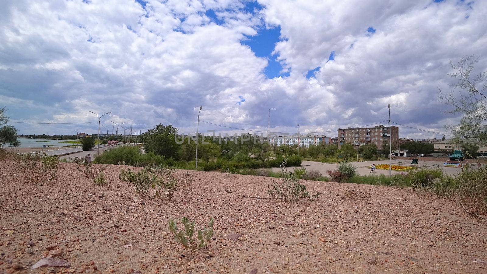 A small town with big clouds and a factory. Alley, green trees swaying in the wind, flowers in flower beds. People are walking. In the distance you can see a lake, reeds grow. Balkhash city