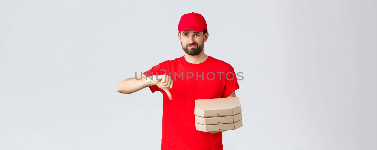 Food delivery, quarantine, stay home and order online concept. Disappointed and reluctant courier in red uniform cap and t-shirt, dislike pooq quality fast-food, thumb-down, holding pizza by Benzoix
