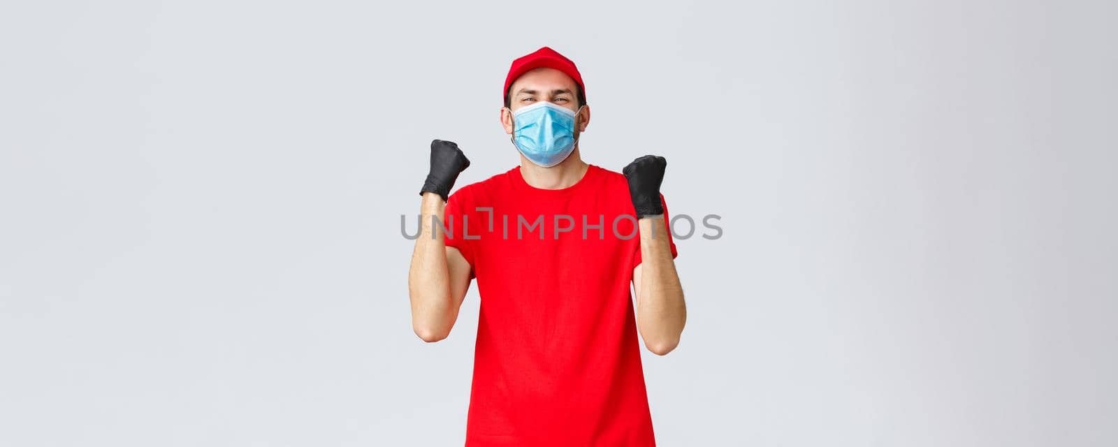Covid-19, self-quarantine, online shopping and shipping concept. Excited, happy courier in red uniform, medical face mask, fist pump in rejoice. Delivery guy celebrating success, achieve goal.