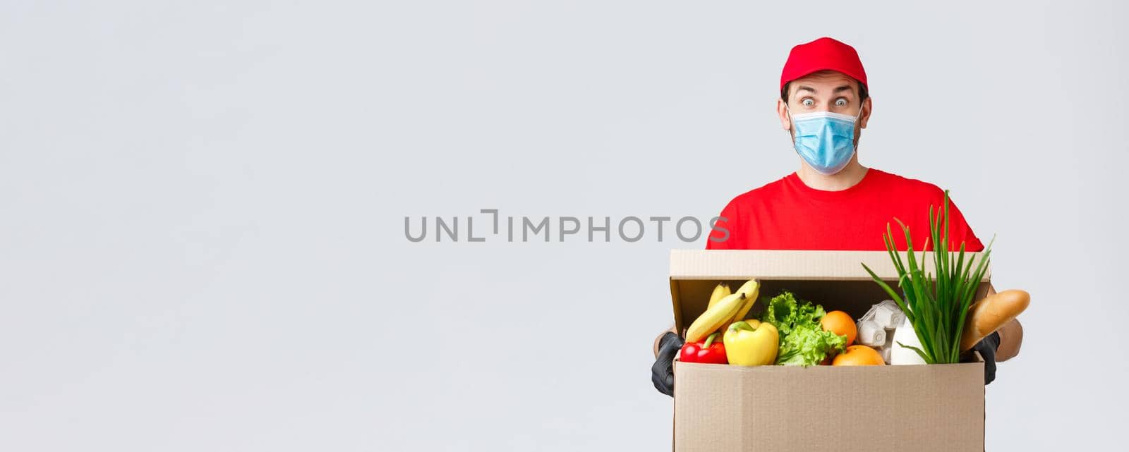 Groceries and packages delivery, covid-19, quarantine and shopping concept. Surprised courier in red uniform, face mask and gloves, impressed, holding box with food, express deliver of client orders by Benzoix