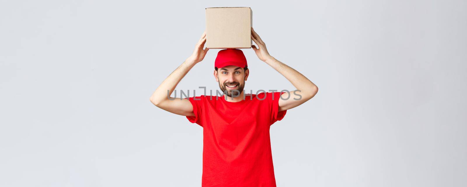 Order delivery, online shopping and package shipping concept. Funny and cute bearded courier in red uniform cap and t-shirt, holding box on head. Employee with package smiling.