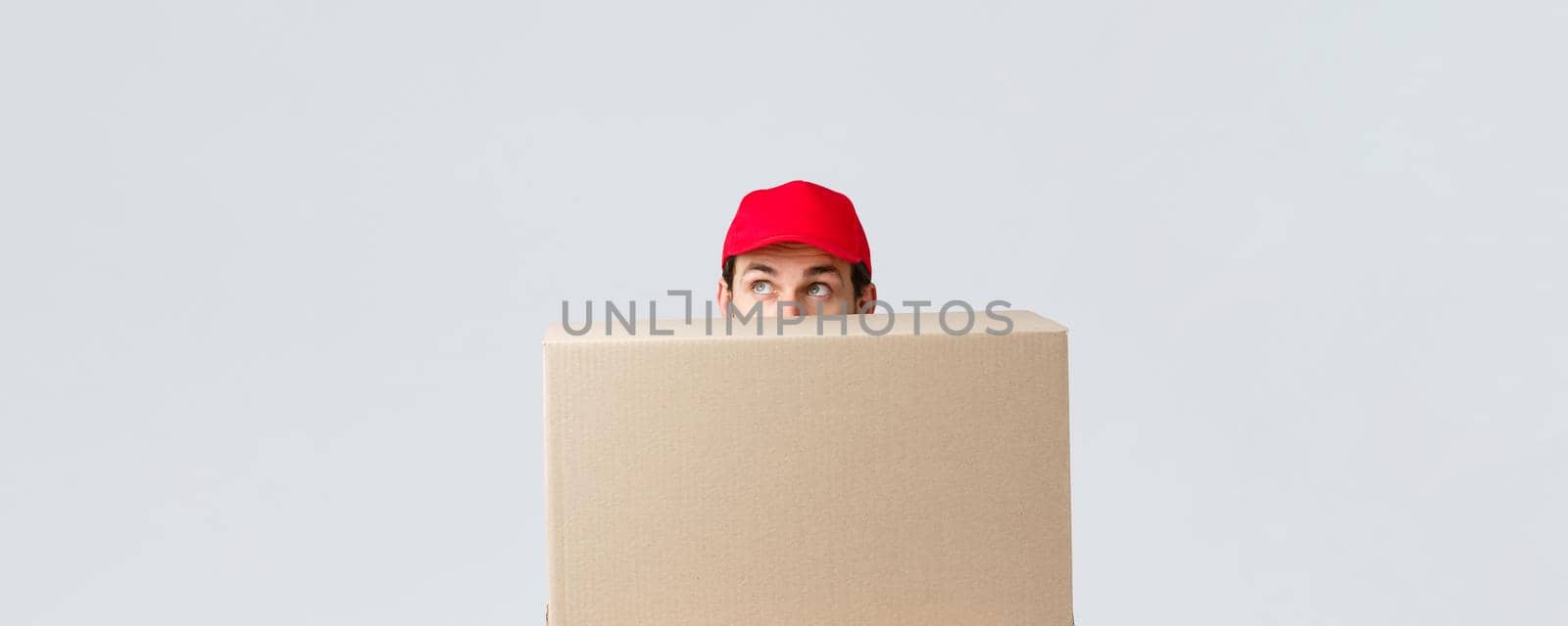 Packages and parcels delivery, covid-19 quarantine and transfer orders. Cute courier in red uniform cap hiding behind large box order, looking up intrigued, peeking over grey background by Benzoix