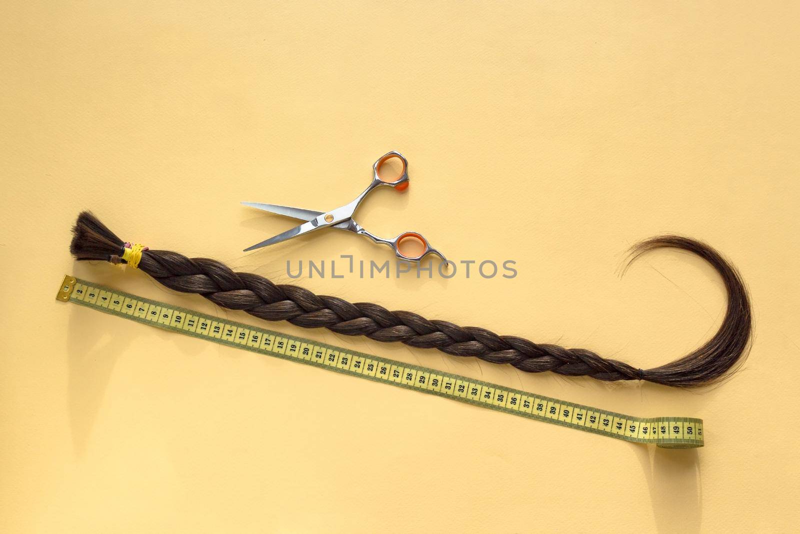 Long brown braid with scissors and tape measure on yellow background, donation of haircut, haircut selling service, material of natural hair for model hairstylish extension, top view