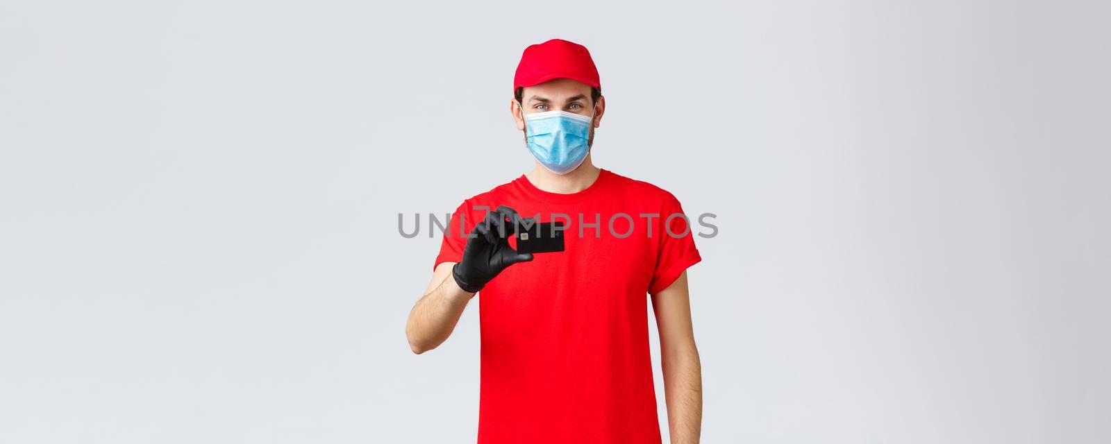 Contactless delivery, payment and online shopping during covid-19, self-quarantine. Handsome courier in red uniform, cap, medical face mask and gloves, show credit card, order internet.