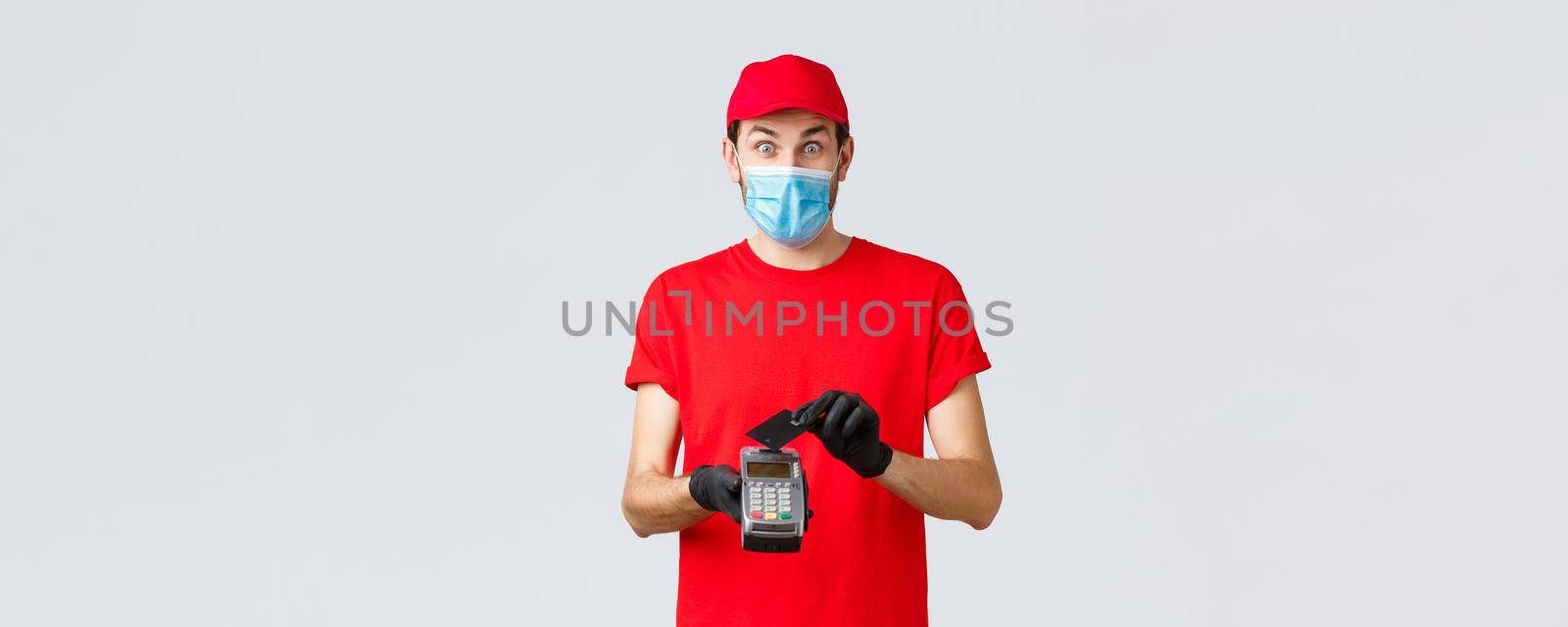 Contactless delivery, payment and online shopping during covid-19, self-quarantine. Excited courier in red uniform, face mask and gloves, look surprised, using credit card on paying terminal POS.