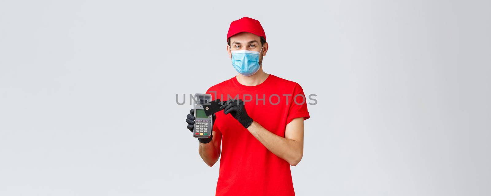 Contactless delivery, payment and online shopping during covid-19, self-quarantine. Friendly courier in red uniform, gloves and face mask, showing paying order with POS terminal and credit card by Benzoix