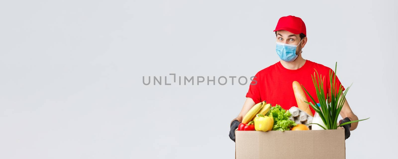 Groceries and packages delivery, covid-19, quarantine and shopping concept. Friendly courier in face mask and gloves, red uniform bring food box to customer ordered online, contactless deliver by Benzoix