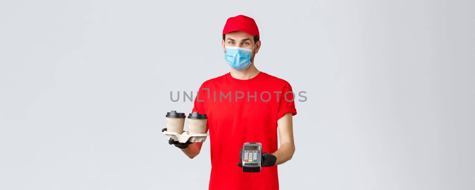 Takeaway, food and groceries delivery, covid-19 contactless orders concept. Pleasant courier in red uniform, gloves and face mask, holding coffee for client and POS terminal, grey background by Benzoix