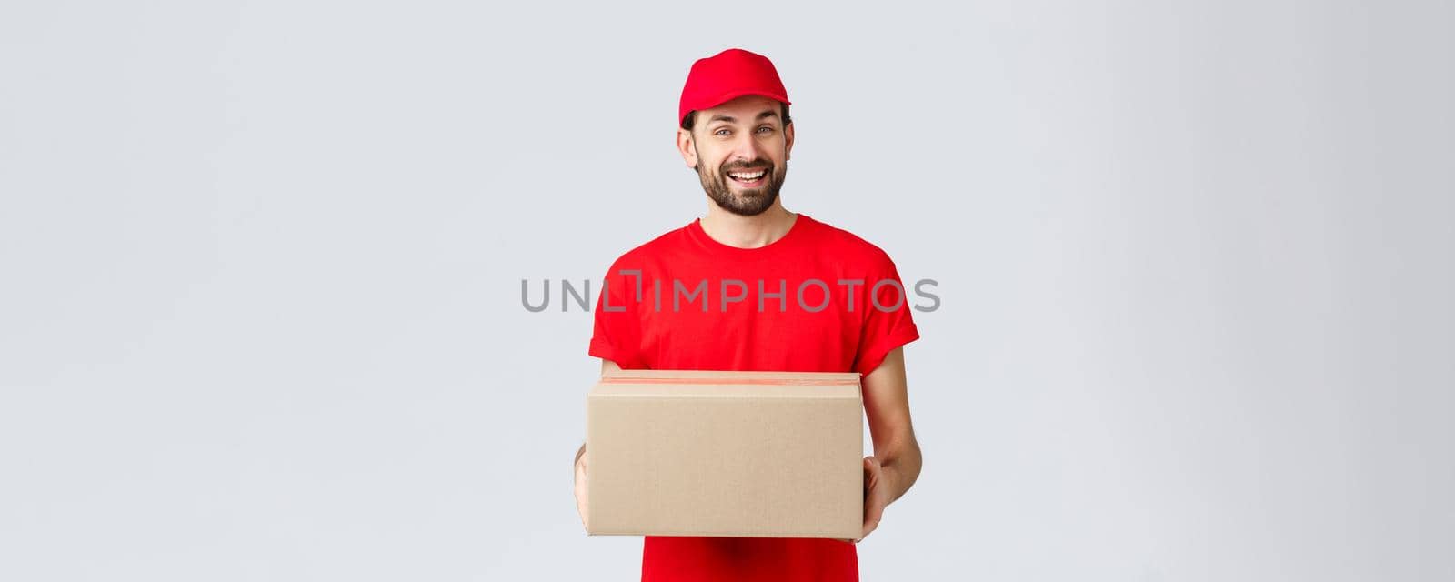 Order delivery, online shopping and package shipping concept. Friendly smiling courier in red uniform cap and t-shirt, handing out packages for customers. Employee bring parcel box, grey background by Benzoix
