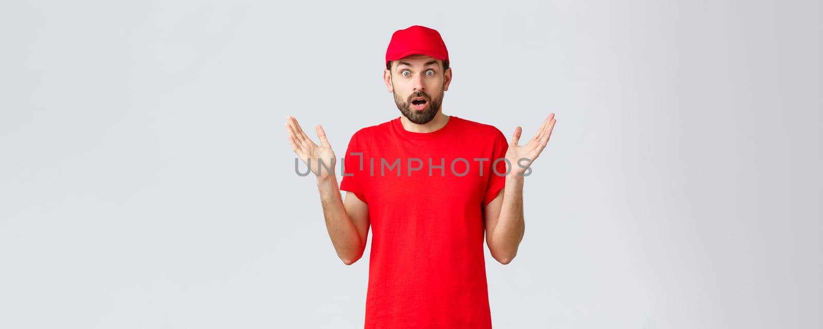 Online shopping, delivery during quarantine and takeaway concept. Confused and shocked courier in red t-shirt and cap of company service, raise hands up indecisive and nervous, cant believe smth by Benzoix