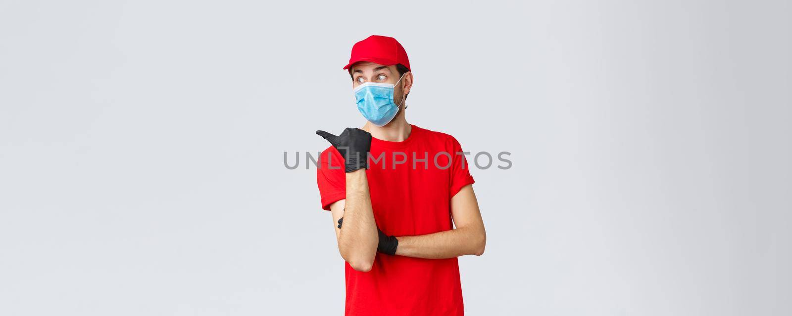 Covid-19, self-quarantine, online shopping and shipping concept. Surprised courier in red uniform, face mask and gloves, pointing thumb and look left impressed, curiously reading promo, show way.