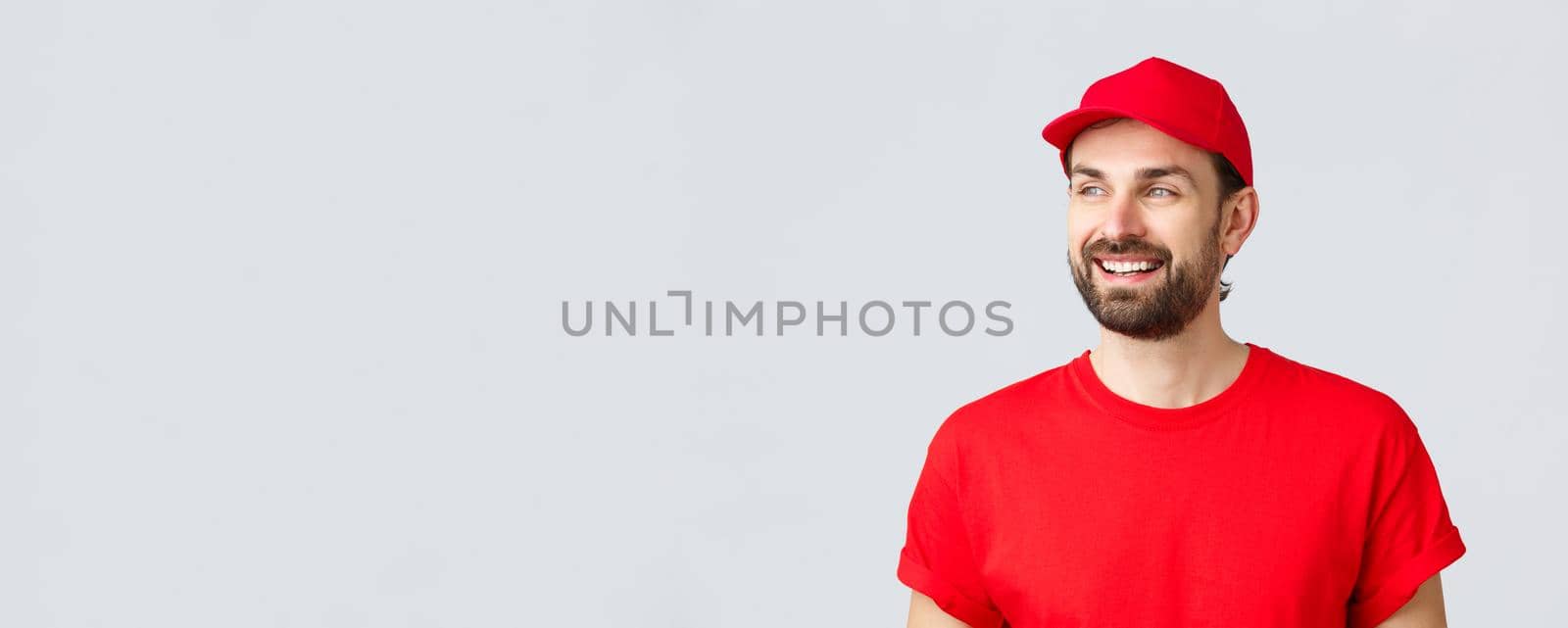 Online shopping, delivery during quarantine and takeaway concept. Cheerful bearded guy in red uniform cap and t-shirt, looking away with pleased smile, reading banner sign, grey background.