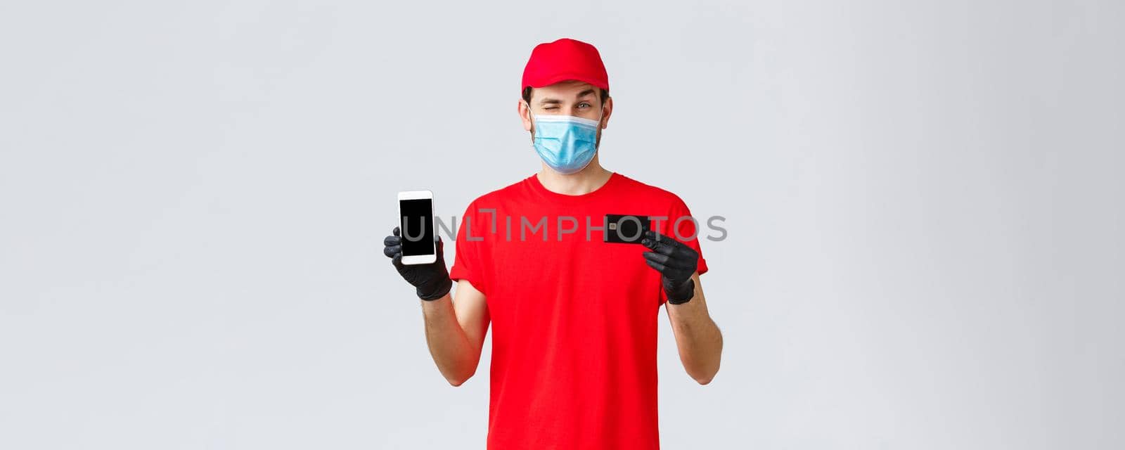 Contactless delivery, payment, online shopping during covid-19, self-quarantine. Handsome courier in red uniform, gloves and face mask, showing smartphone screen and credit card, promo of order app.