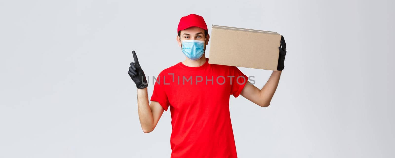 Packages and parcels delivery, covid-19 quarantine delivery, transfer orders. Cheerful courier in face mask, gloves and uniform, carry your parcel to doorsteps, pointing up, hold box with order.