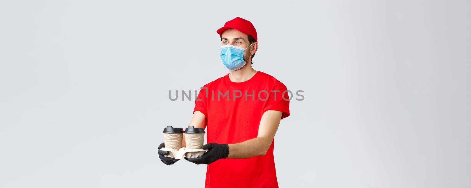 Takeaway, food and groceries delivery, covid-19 contactless orders concept. Pleasant courier in red uniform, face mask and gloves, giving out coffee to customer, stand grey background.