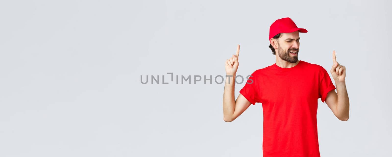 Upbeat handsome bearded delivery guy spending break relaxing, employee in red uniform cap and t-shirt singing song, pointing fingers up carefree, close eyes, standing grey background.
