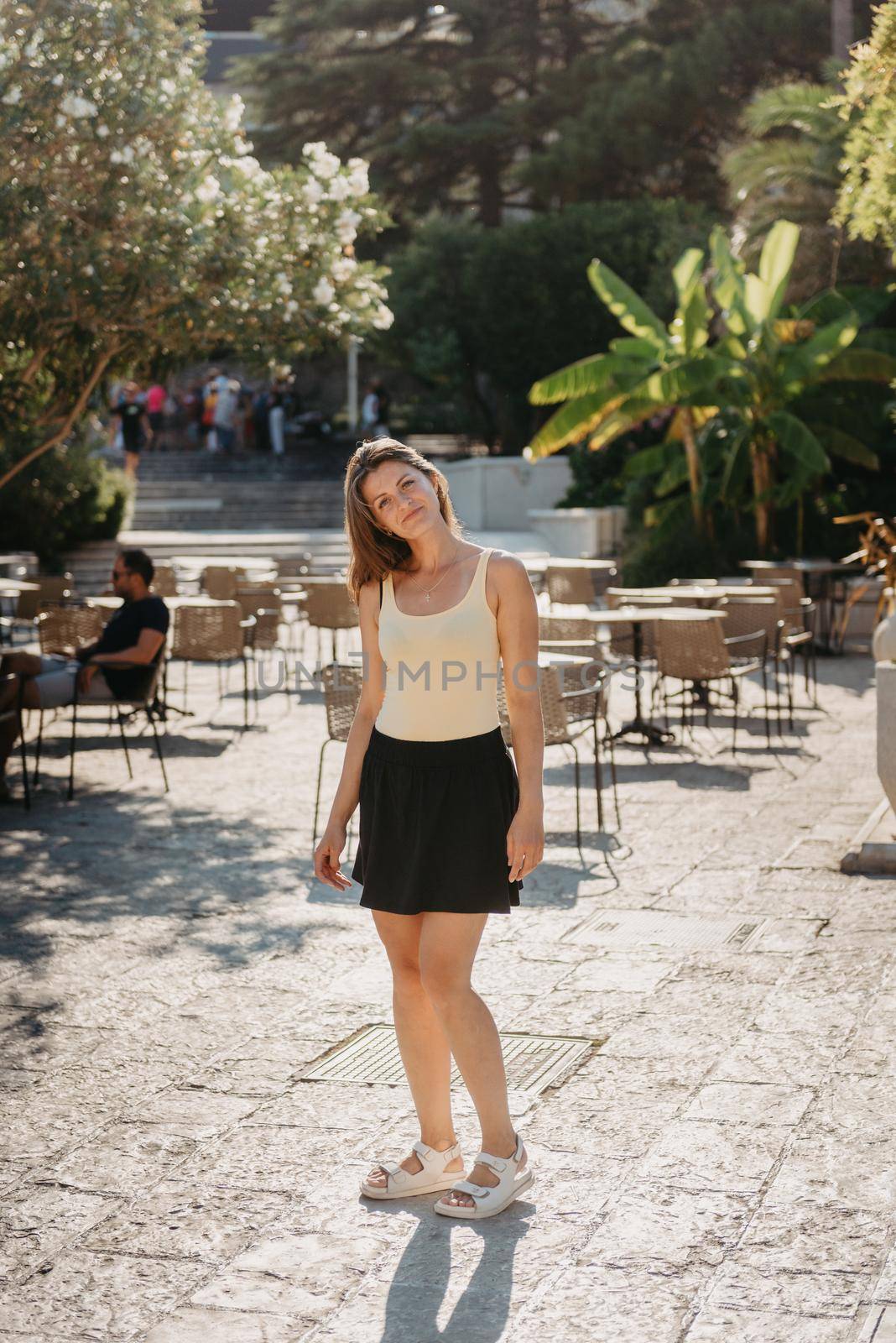 Girl tourist walking through ancient narrow street on a beautiful summer day in MEDITERRANEAN MEDIEVAL CITY , OLD TOWN bUDVA, MONTENEGRO. Young beautiful cheerful woman walking on old street at tropical town. Pretty girl looking at you and smiling