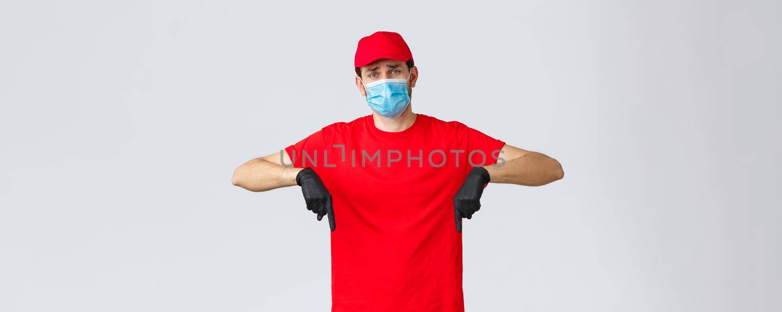 Covid-19, self-quarantine, online shopping and shipping concept. Upset and gloomy, sad delivery guy in red cap, t-shirt uniform, pointing fingers down disappointed. Courier showing bad news.
