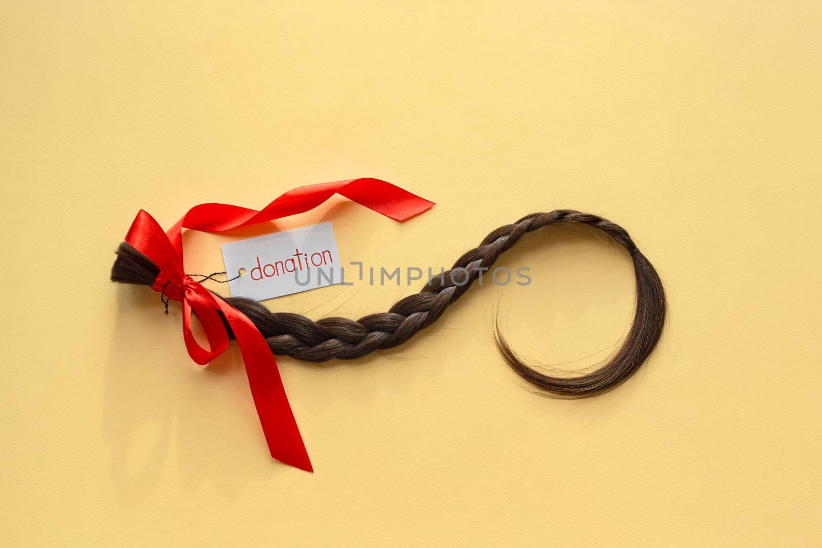 Long brown braid with tag label and red ribbon on yellow background, donation of haircut, natural human female hair, charity support and acts of kindess, top view