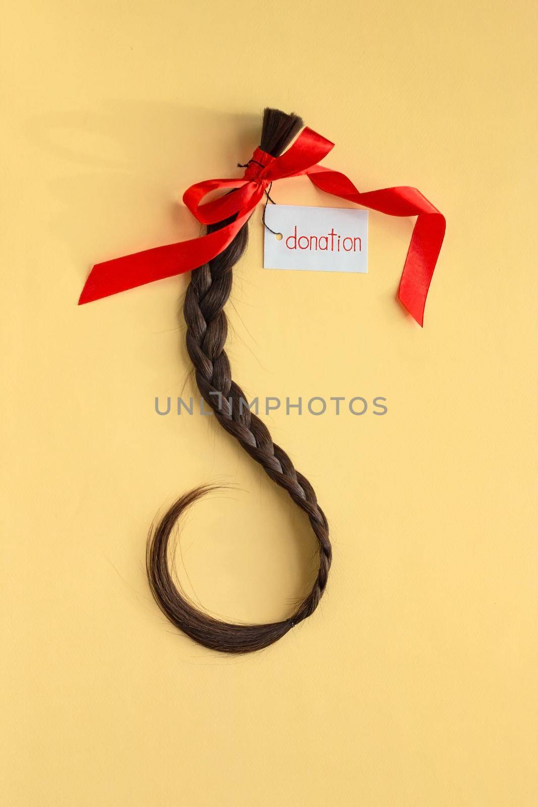 Long brown braid with tag label and red ribbon on yellow background, donation of haircut, natural human female hair, charity support and acts of kindess, top view, vertical image