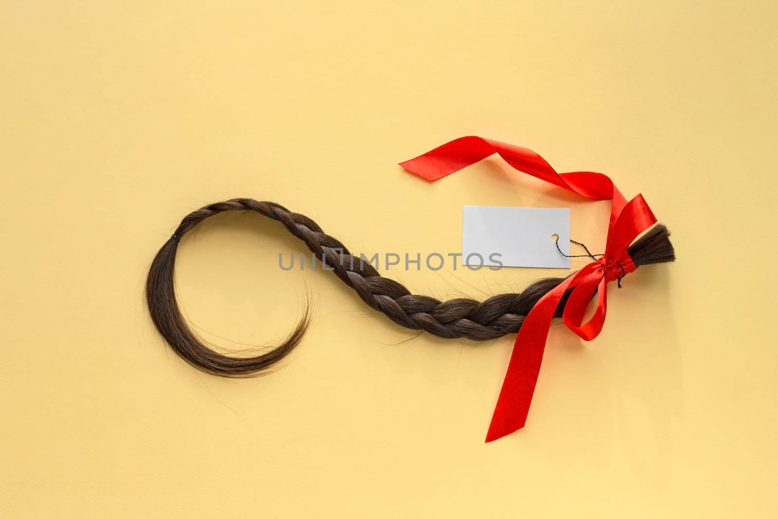 Long brown braid with red ribbon and empty label on yellow background, haircut selling service, material of natural hair for model hairstylish extension, top view
