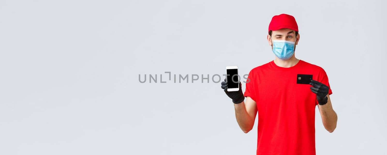 Contactless delivery, payment, online shopping during covid-19, self-quarantine. Handsome courier in red uniform, gloves and face mask, showing smartphone screen and credit card, promo of order app by Benzoix