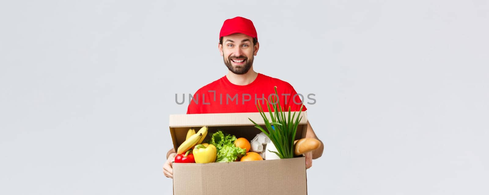 Groceries and packages delivery, covid-19, quarantine and shopping concept. Smiling delivery man in red uniform, bring client order, holding food box parcel, enjoying help people transfer goods by Benzoix