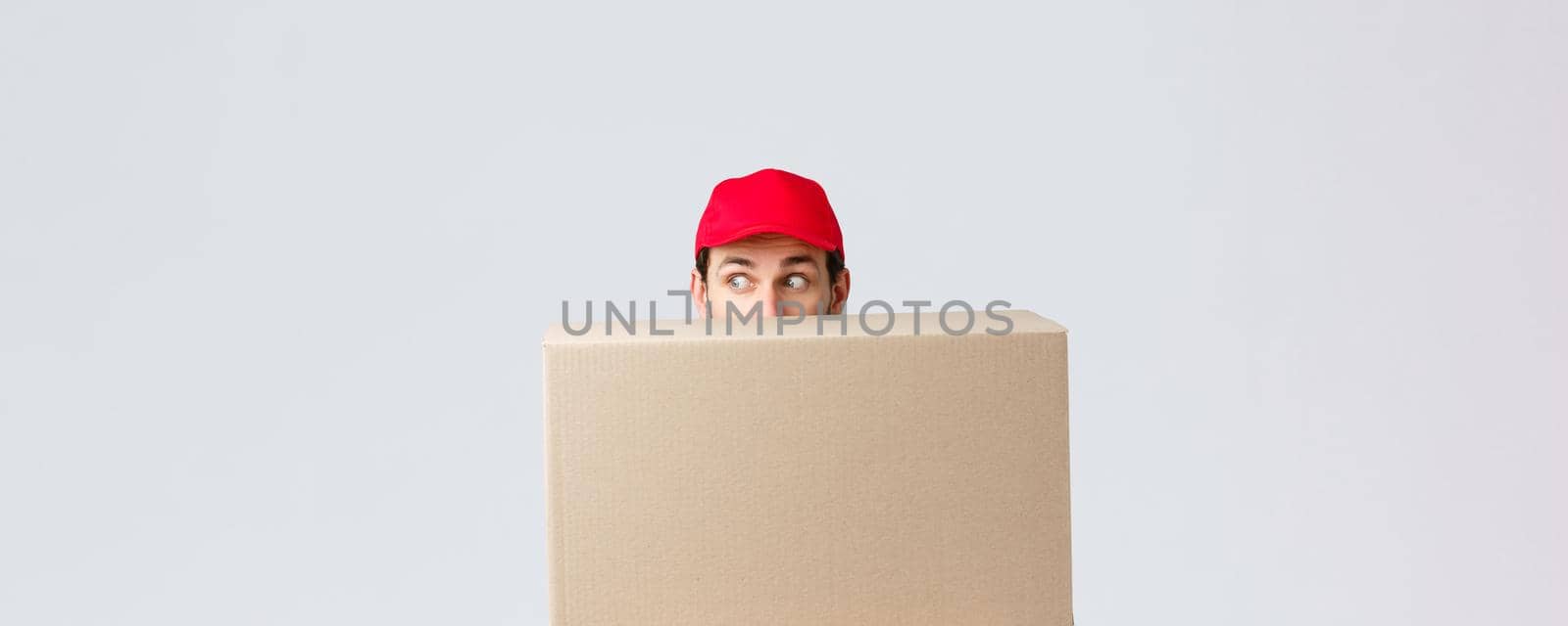 Packages and parcels delivery, covid-19 quarantine and transfer orders. Scared courier in red uniform cap, hiding behind customer order, looking left nervously, peeking at banner or advertisement by Benzoix