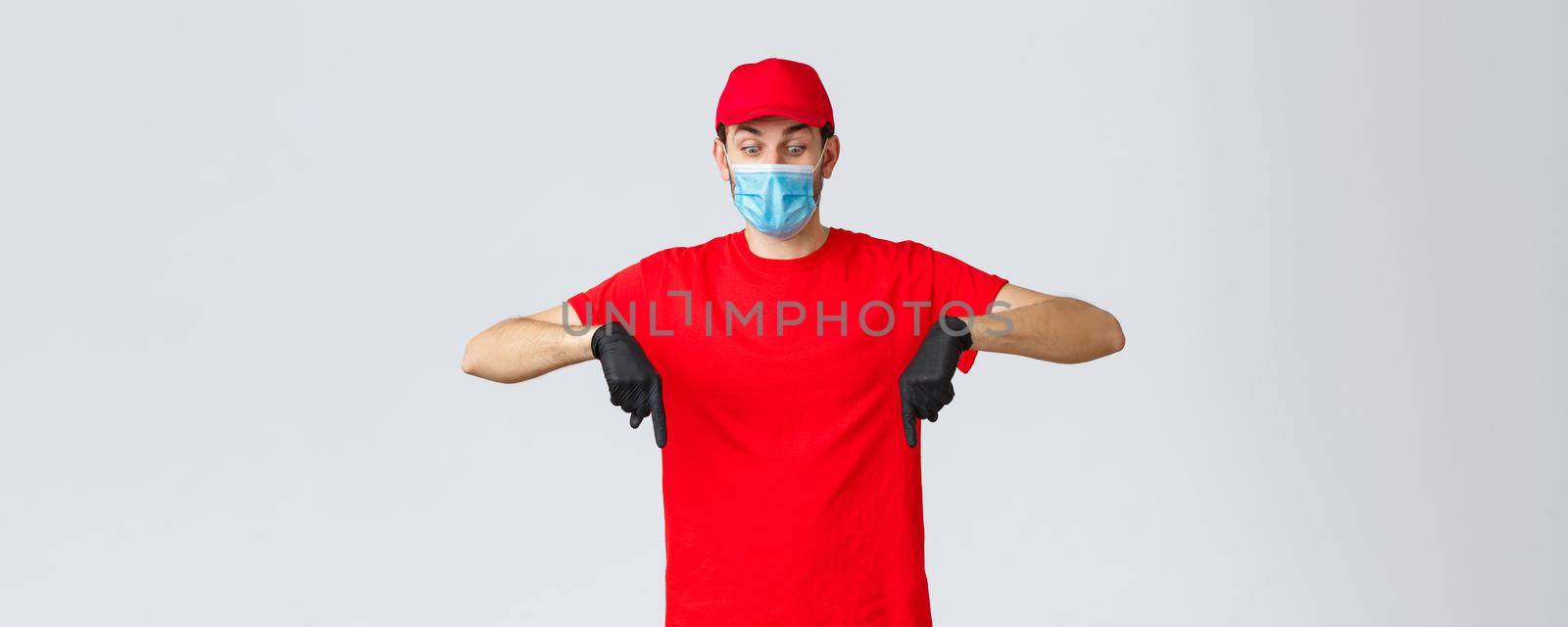 Covid-19, self-quarantine, online shopping and shipping concept. Startled and shocked courier in uniform, red cap and t-shirt, wearing safety measures medical mask and gloves, pointing down.