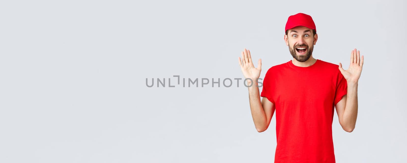 Online shopping, delivery during quarantine and takeaway concept. Happy cheerful courier in red t-shirt and cap, company uniform, hands up surprised and amused, standing grey background.