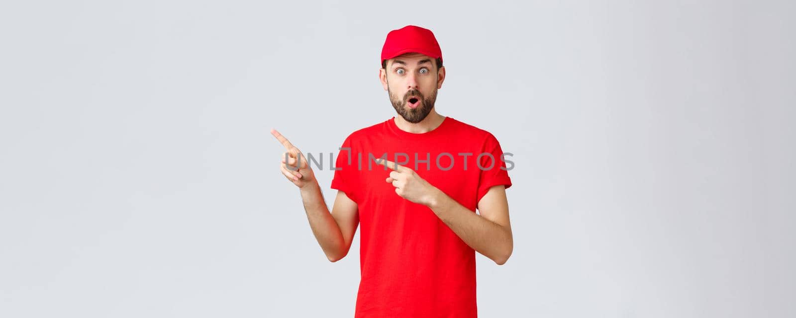 Online shopping, delivery during quarantine and takeaway concept. Shocked and impressed, gasping courier in red t-shirt and cap, uniform of service company, pointing fingers left astonished.
