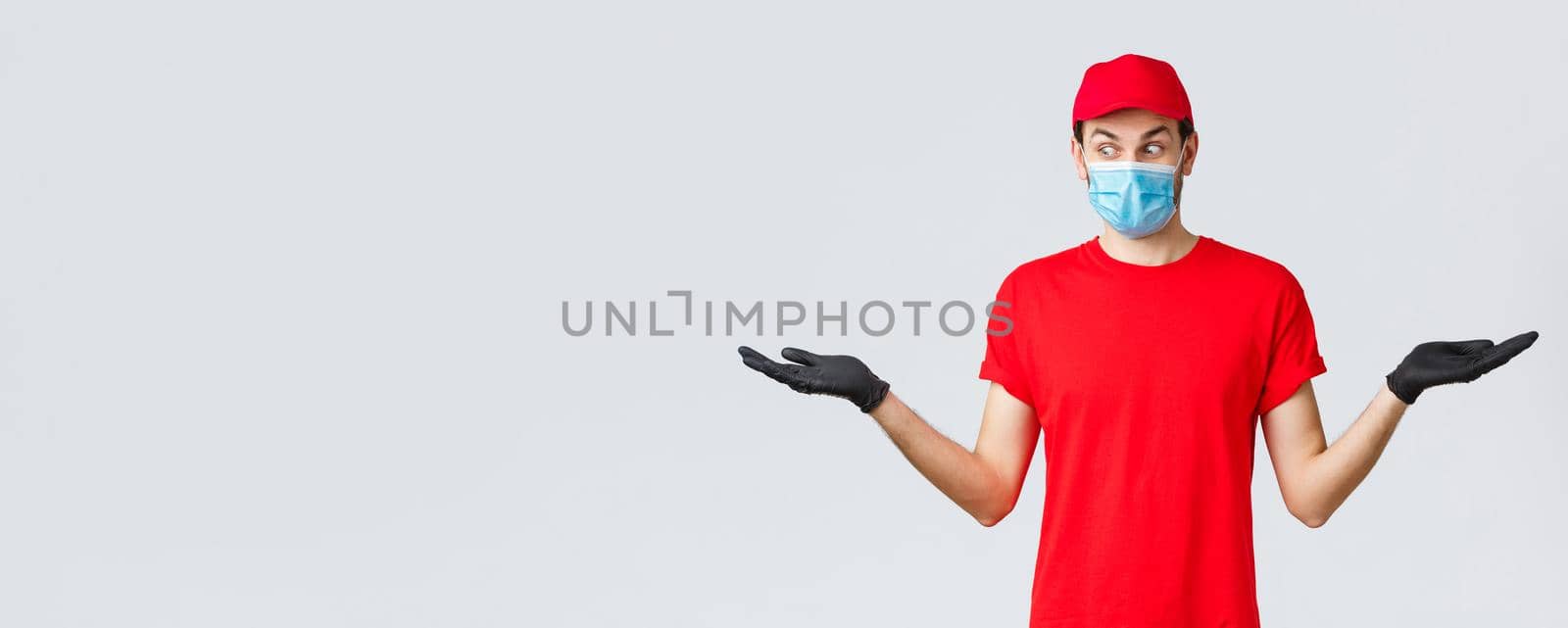 Groceries and packages delivery, covid-19, quarantine and shopping concept. Surprised courier in red cap and t-shirt uniform, wear protective face mask, gloves, hold two items in hands sideways by Benzoix