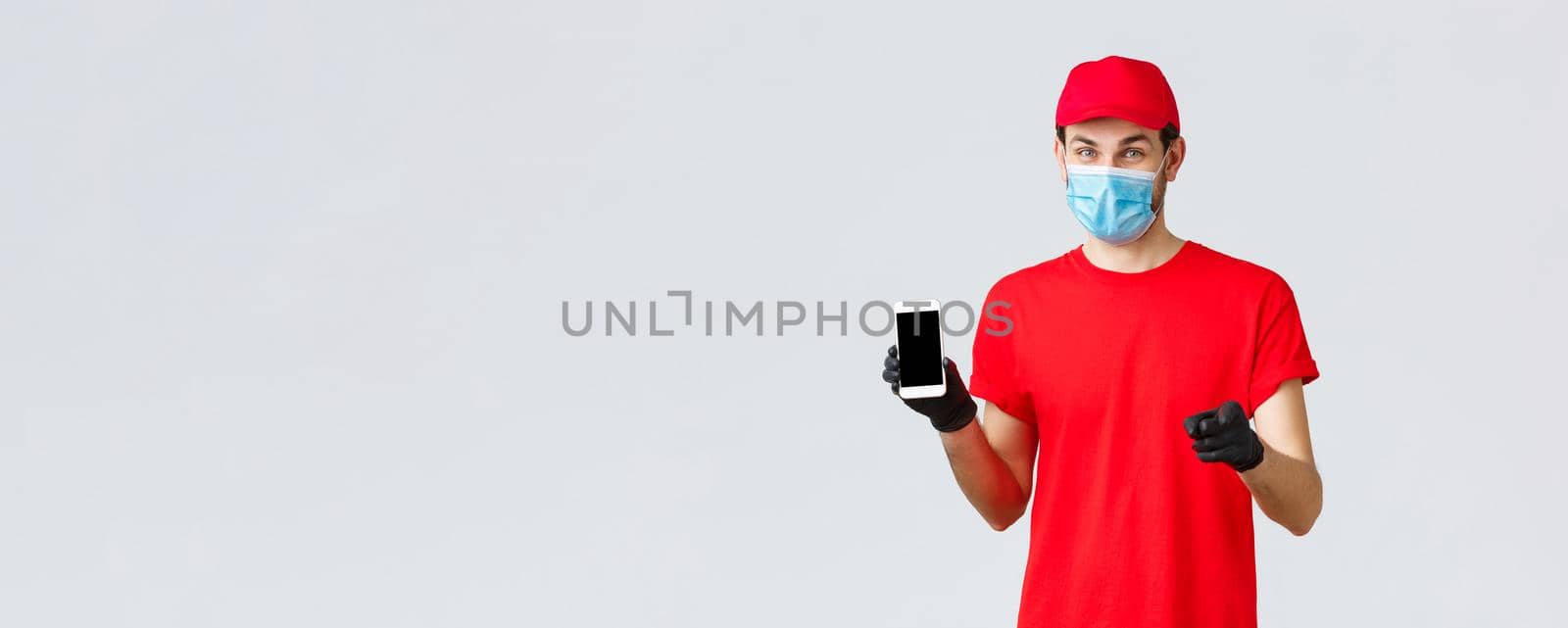 Contactless delivery, payment and online shopping during covid-19, self-quarantine. Smiling courier in red uniform, gloves and face mask, showing order info on phone screen, pointing camera by Benzoix