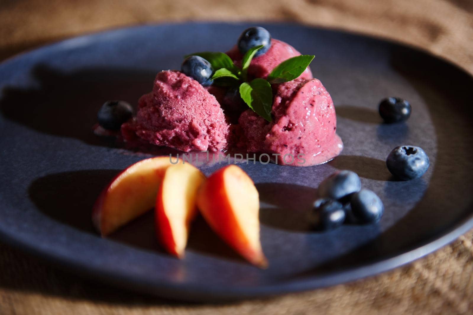 Food still life. Fresh delicious vegan ice cream of ripe organic blueberries, melting on a navy blue plate, decorated with mint and lemon basil leaves, ripe juicy peaches. Healthy homemade sorbet