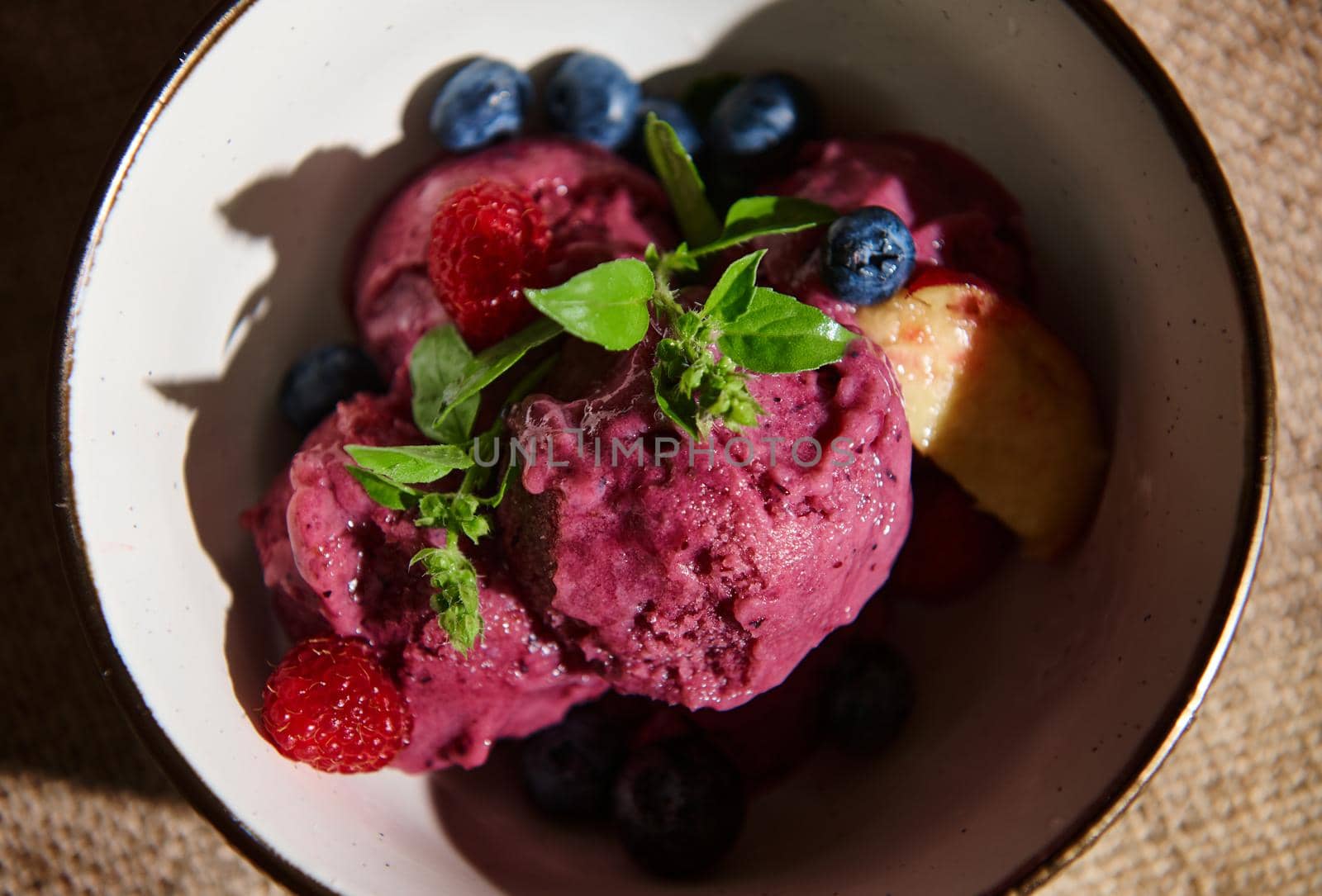 Flat lay. Food still life. Homemade purple, raw vegan, healthy berry ice cream sorbet, with ripe juicy fruits, blueberries and lemon basil leaves in a ceramic bowl on a linen tablecloth. Copy ad space