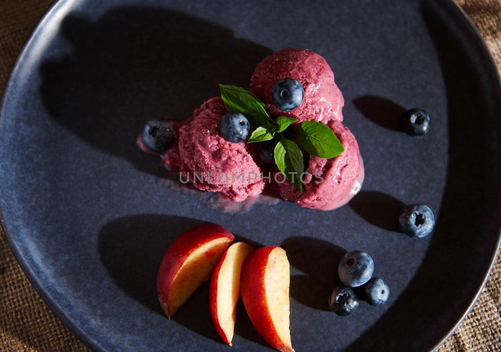 Food still life. Refreshing raw vegan huckleberry ice cream sorbet, melting on a navy blue plate, decorated with mint, lemon basil leaves, blueberries and ripe juicy peaches. Healthy homemade dessert