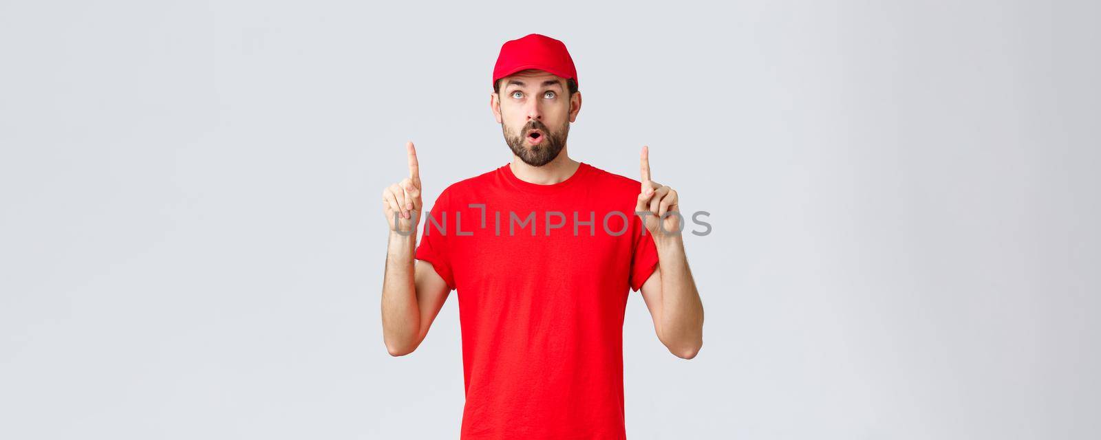 Online shopping, delivery during quarantine and takeaway concept. Impressed, curious employee in red uniform cap and t-shirt, open mouth interested, read banner, pointing fingers up by Benzoix