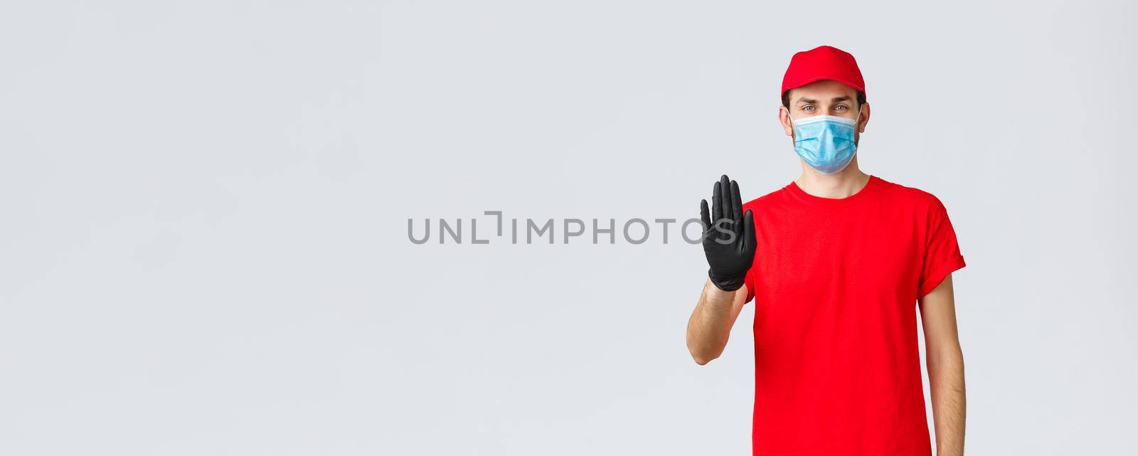 Covid-19, self-quarantine, online shopping concept. Confident delivery man in red uniform, gloves and face mask, raise hand in stop sign, prevent client step to storage without protective equipment by Benzoix