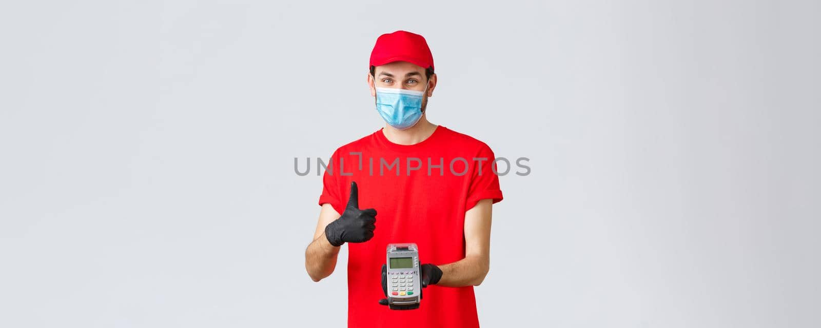 Contactless delivery, payment and online shopping during covid-19, self-quarantine. Friendly smiling courier in red uniform cap, t-shrit, medical mask and gloves, advice pay order with POS terminal by Benzoix