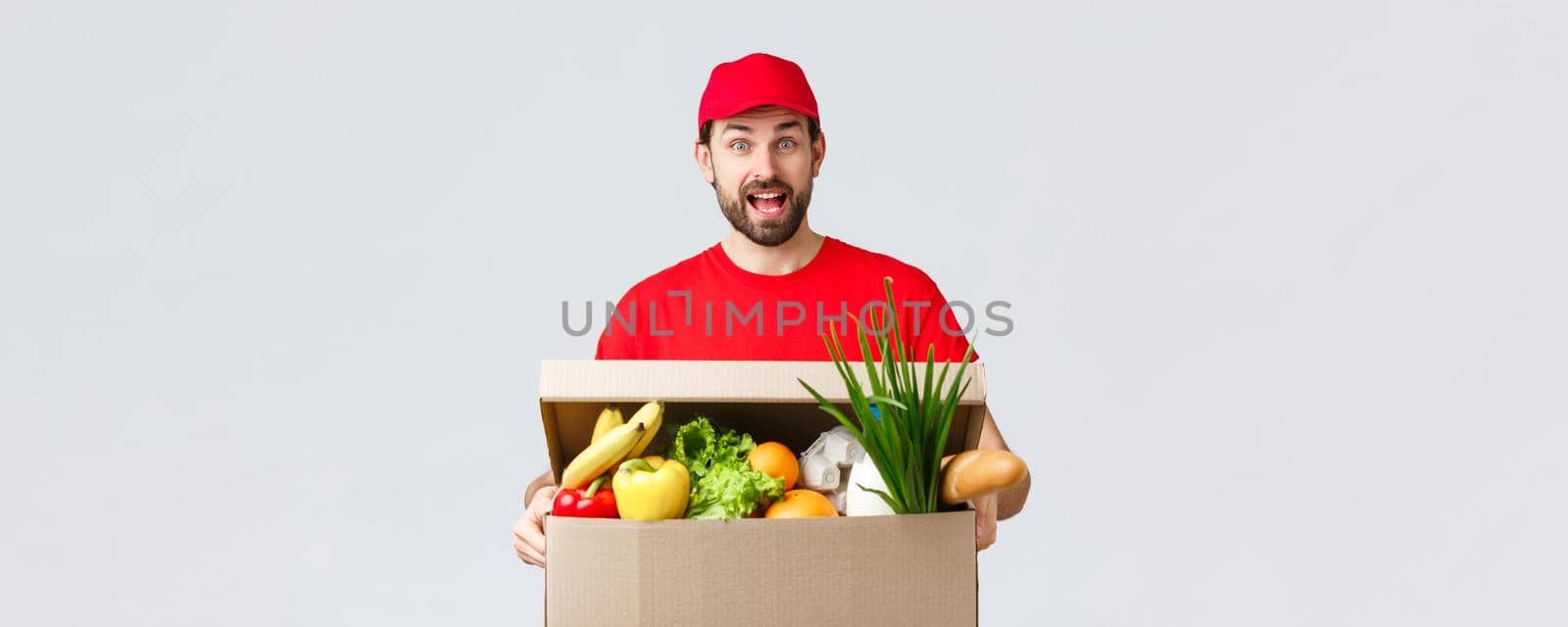 Groceries and packages delivery, covid-19, quarantine and shopping concept. Smiling handsome bearded courier in red uniform, bring food package, grocery order to client in box, look amused by Benzoix